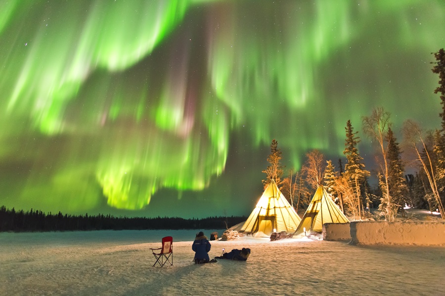 Das Bild zeigt eine Winterlandschaft im Norden Kanadas, über der Polarlichter zu sehen sind. Sie ziehen sich wie im Wind wehende Vorhänge über den gesamten Nachthimmel. Im Vordergrund kniet ein Mensch neben zwei erleuchteten Zelten.